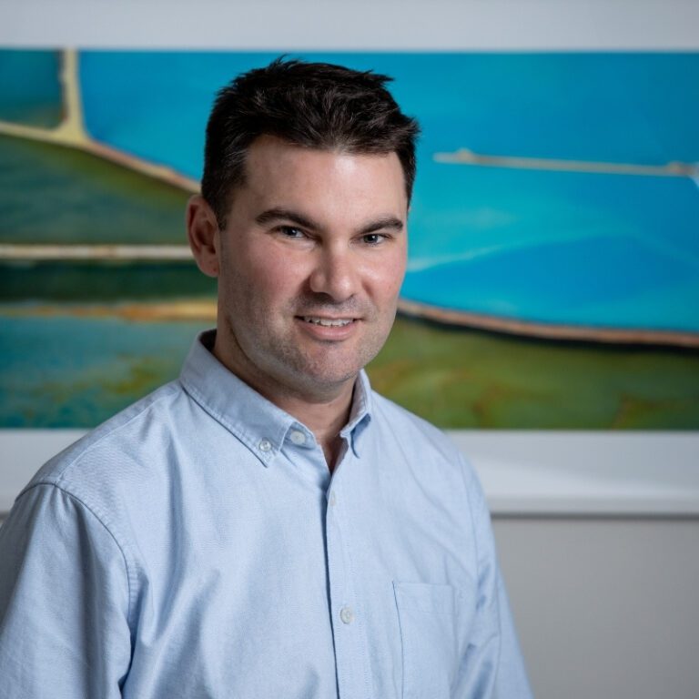 Headshot of a smiling male psychiatrist, Dr. Luke Somerville-Brown, wearing a light blue button-up shirt in front of a blue and turquoise painting.